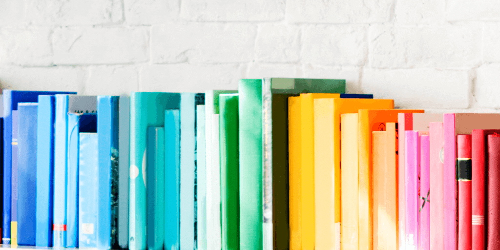 Books arranged on a shelf in a gradient of colors from blue to green, yellow, orange, and red evoke the visual appeal of 21 Instagram statistics marketers really need, set against a crisp white brick wall background.