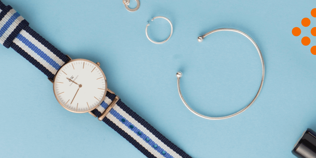A striped wristwatch, silver bracelet, two silver rings, and part of a black rectangular object are arranged on a light blue background with small orange dots in the corner—perfect to promote your products with rich pins on Pinterest.