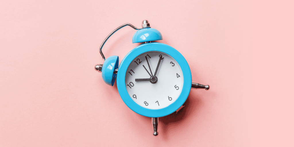 A blue retro alarm clock with a bent bell lies on its side against a pastel pink background, reminding you it’s almost time to try 9 low cost productivity automation tools. The clock shows the time as 11:55.